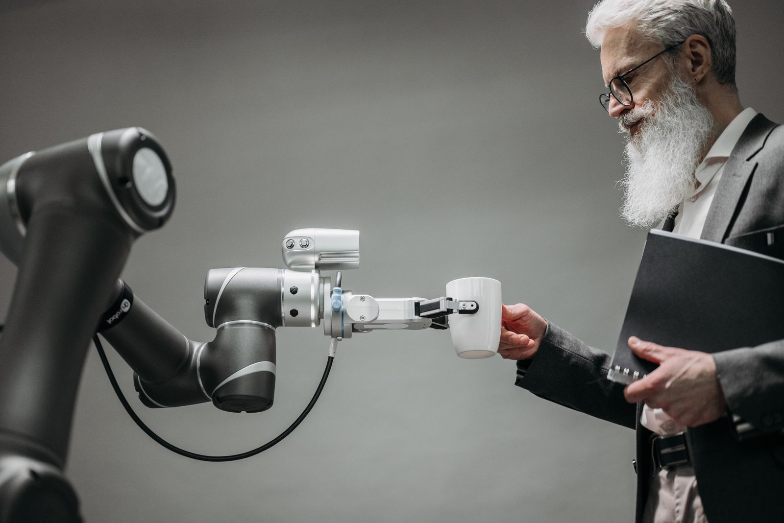 A robot arm hands a white mug to a bearded man on a grey background — human-AI interaction