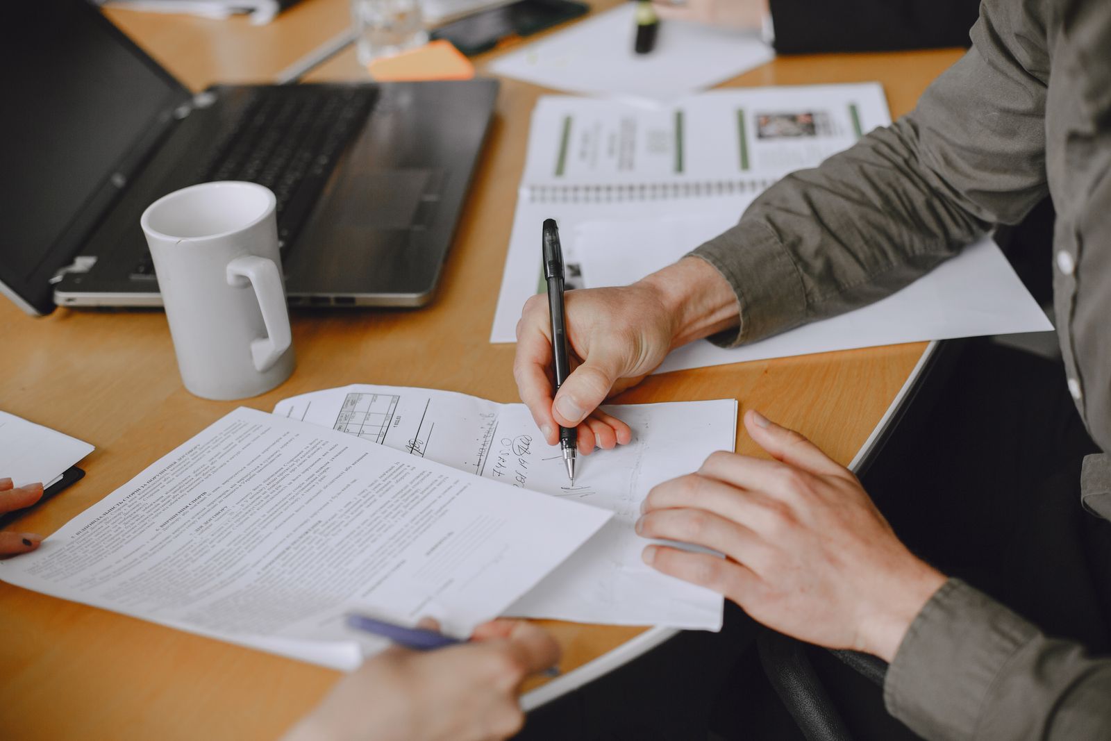 A person signing a printed document at a desk next to a laptop and a mug