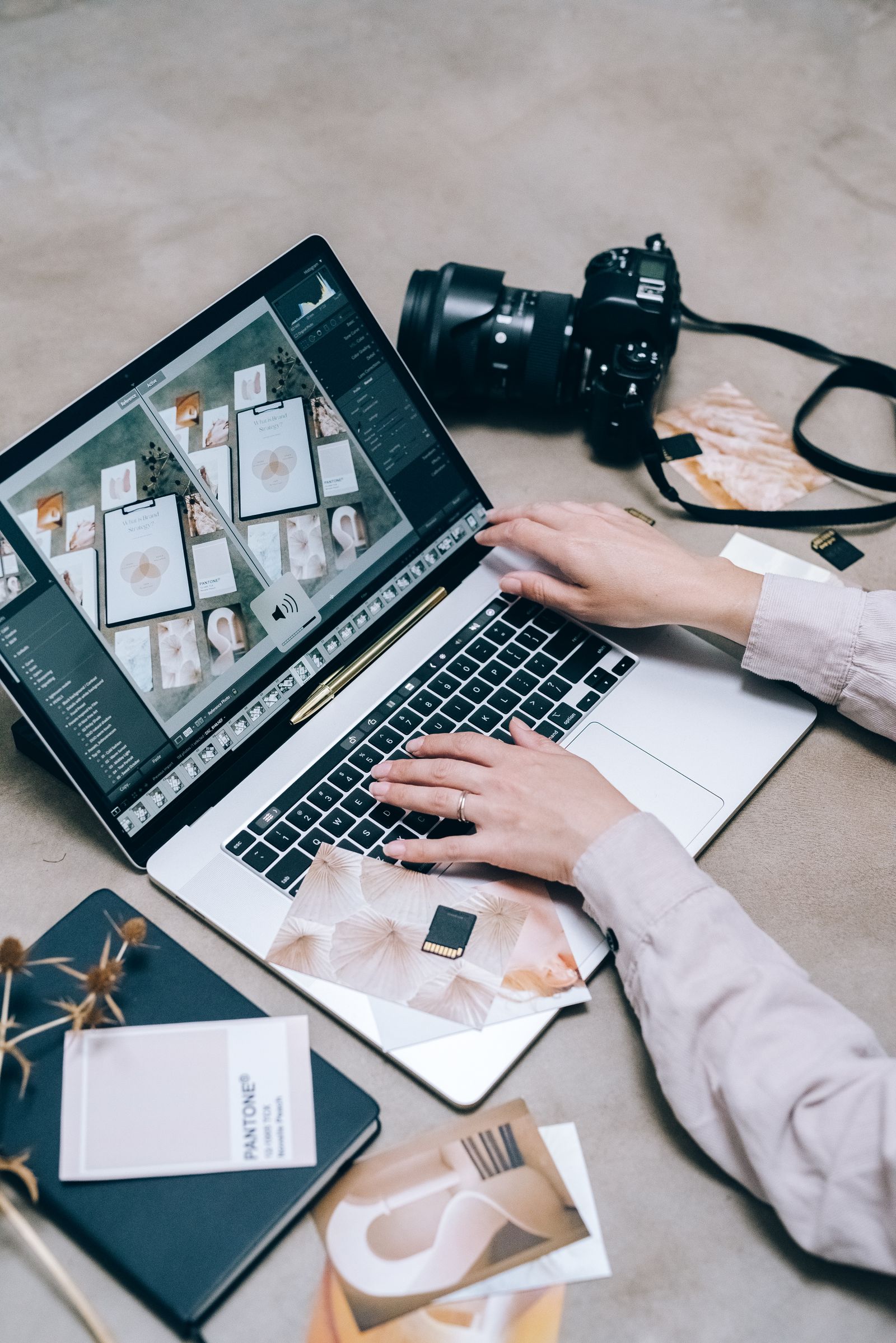 Hands typing on a laptop with an editor open, a camera and a Pantone swatch book nearby